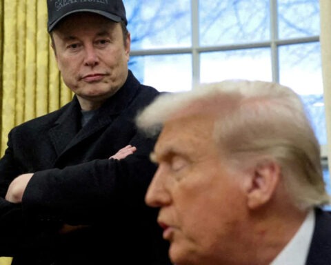 Elon Musk watches Donald Trump speak during a White House meeting, wearing a cryptic expression and a hat that reads “Great Again”