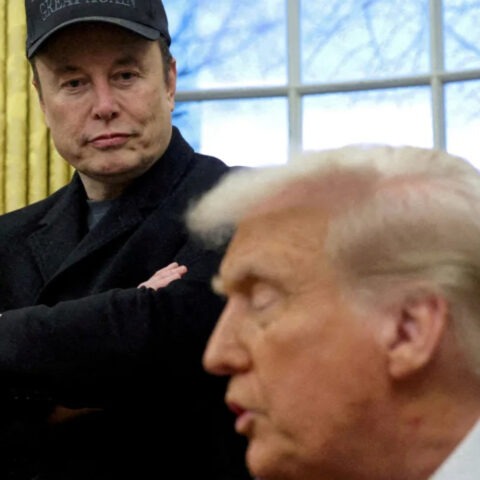 Elon Musk watches Donald Trump speak during a White House meeting, wearing a cryptic expression and a hat that reads “Great Again”