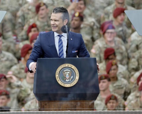 Pete Hegseth speaks at podium with the presidential seal, surrounded by troops in camouflage uniforms, during a military event supporting Trump’s base renaming plan