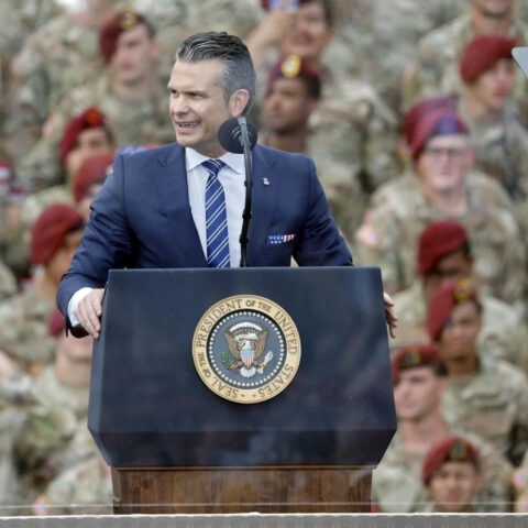 Pete Hegseth speaks at podium with the presidential seal, surrounded by troops in camouflage uniforms, during a military event supporting Trump’s base renaming plan