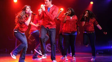 Cast members in red jackets perform on stage under dramatic lighting during a musical number