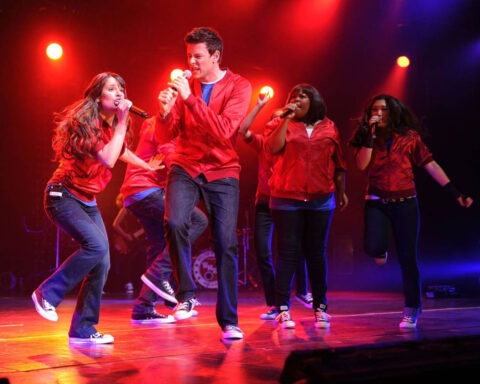 Cast members in red jackets perform on stage under dramatic lighting during a musical number