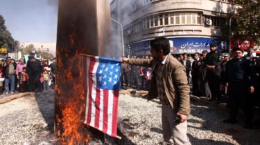 A Middle Eastern merchant surrounded by people in the street as he burns American flags labeled “flammable”