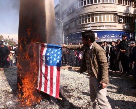 A Middle Eastern merchant surrounded by people in the street as he burns American flags labeled “flammable”