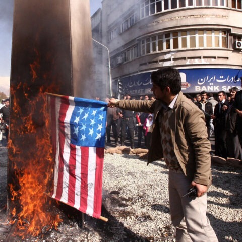 A Middle Eastern merchant surrounded by people in the street as he burns American flags labeled “flammable”