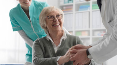 Smiling senior woman in wheelchair shakes hands with a doctor during a checkup, with a nurse standing behind her
