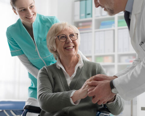 Smiling senior woman in wheelchair shakes hands with a doctor during a checkup, with a nurse standing behind her