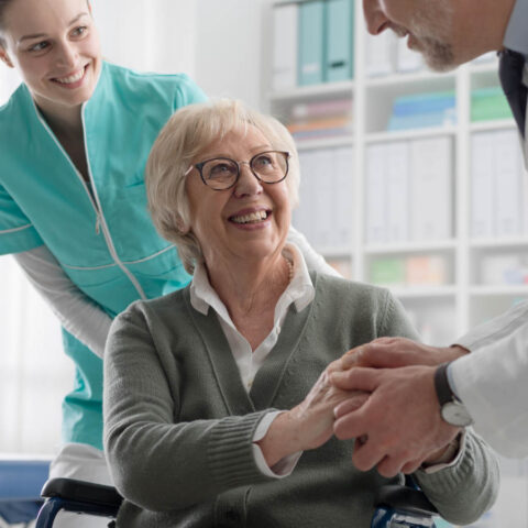 Smiling senior woman in wheelchair shakes hands with a doctor during a checkup, with a nurse standing behind her