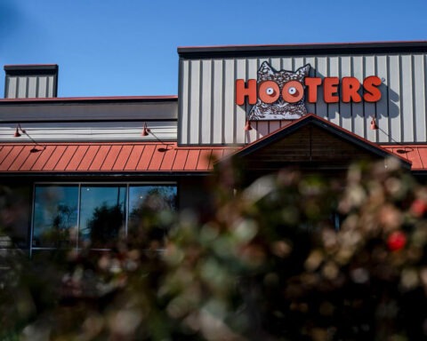 Hooters restaurant with its iconic orange sign seen from behind some bushes, as the chain closes 30 locations nationwide due to shifting family dining preferences.