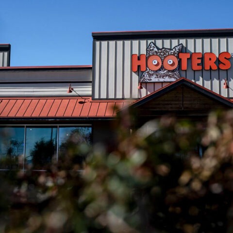 Hooters restaurant with its iconic orange sign seen from behind some bushes, as the chain closes 30 locations nationwide due to shifting family dining preferences.