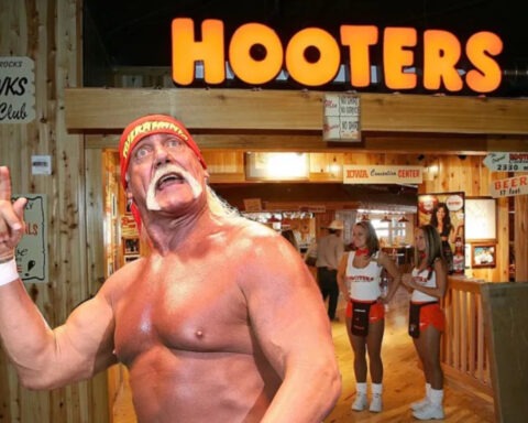 Hulk Hogan poses shirtless in front of a Hooters restaurant while flexing, with two Hooters waitresses in uniform standing in the background