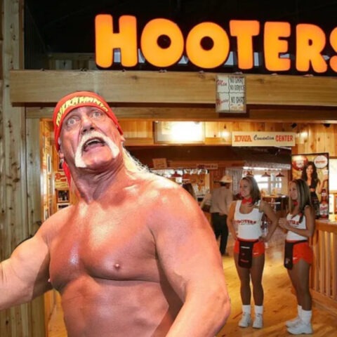 Hulk Hogan poses shirtless in front of a Hooters restaurant while flexing, with two Hooters waitresses in uniform standing in the background