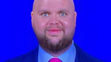 Close-up of a bald man with a trimmed beard and a slightly unsettling smile in front of a bright blue background