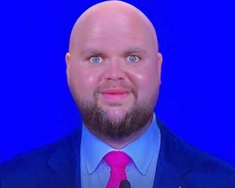 Close-up of a bald man with a trimmed beard and a slightly unsettling smile in front of a bright blue background