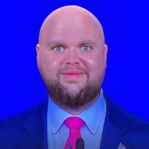 Close-up of a bald man with a trimmed beard and a slightly unsettling smile in front of a bright blue background