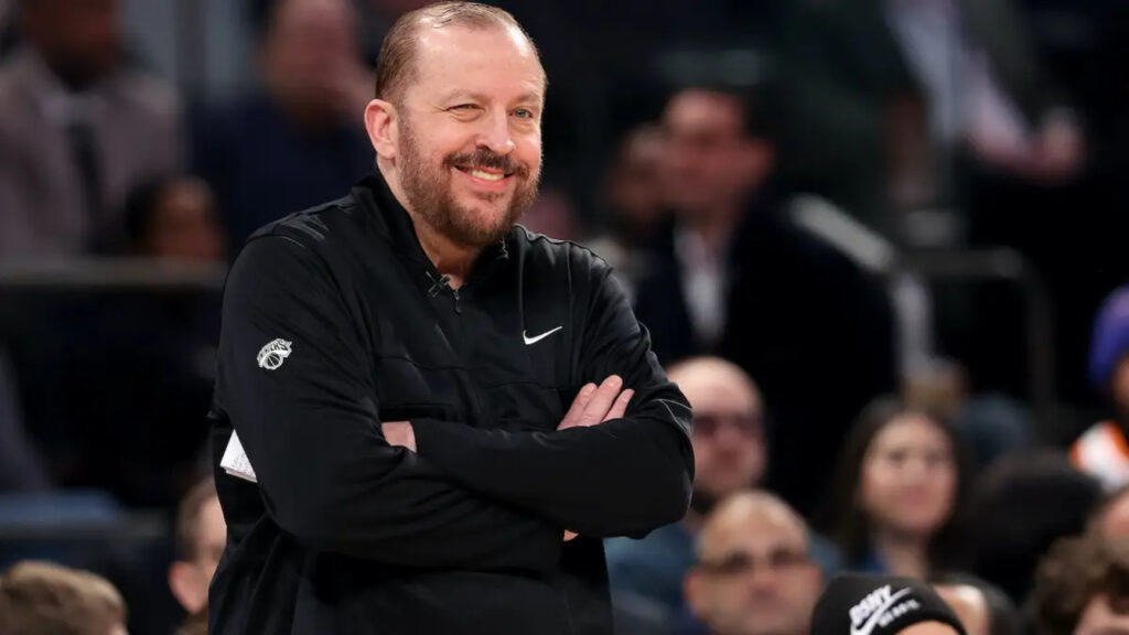 New York Knicks coach Tom Thibodeau winks while smiling courtside during an NBA game.