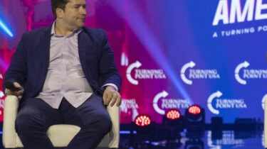 Man in blue suit holding microphone at Turning Point USA event, sitting on stage with colorful background