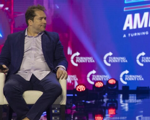 Man in blue suit holding microphone at Turning Point USA event, sitting on stage with colorful background