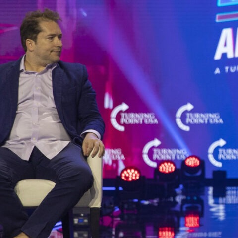 Man in blue suit holding microphone at Turning Point USA event, sitting on stage with colorful background