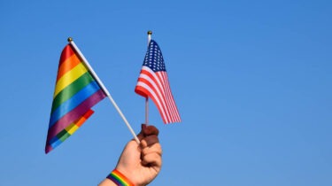 Hand holding a rainbow Pride flag and a U.S. flag against a blue sky