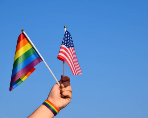 Hand holding a rainbow Pride flag and a U.S. flag against a blue sky