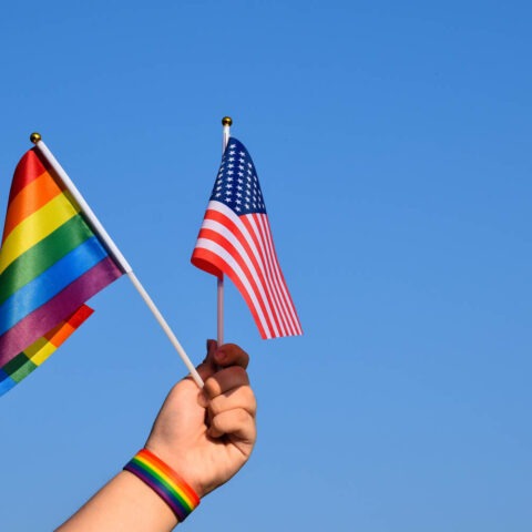 Hand holding a rainbow Pride flag and a U.S. flag against a blue sky