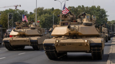 Military tanks with American flags roll down a city street as part of a parade.