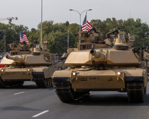 Military tanks with American flags roll down a city street as part of a parade.