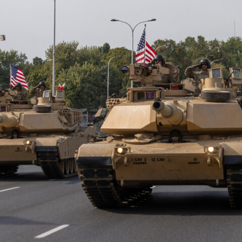 Military tanks with American flags roll down a city street as part of a parade.