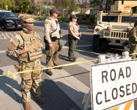 National Guard and sheriff deputies stand at a roadblock with a “Road Closed” sign and military vehicles in a suburban neighborhood
