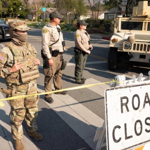 National Guard and sheriff deputies stand at a roadblock with a “Road Closed” sign and military vehicles in a suburban neighborhood