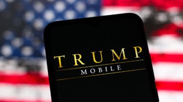 Trump Mobile logo displayed on phone screen with blurry American flag in background