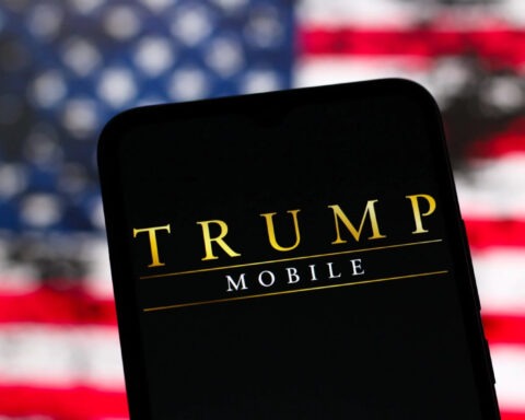 Trump Mobile logo displayed on phone screen with blurry American flag in background