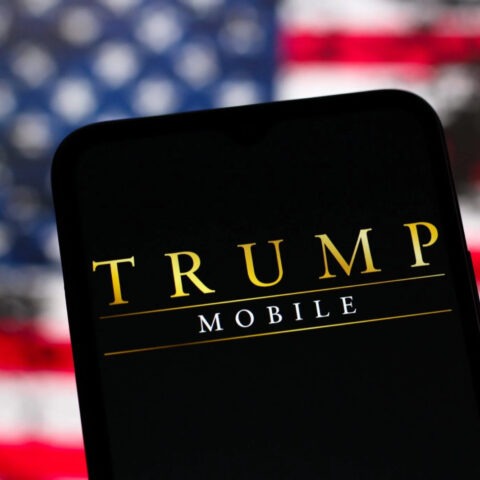 Trump Mobile logo displayed on phone screen with blurry American flag in background
