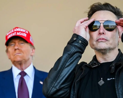 Donald Trump in a red MAGA hat stands behind Elon Musk adjusting his sunglasses, both facing forward with intense expressions.