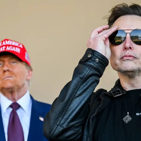 Donald Trump in a red MAGA hat stands behind Elon Musk adjusting his sunglasses, both facing forward with intense expressions.