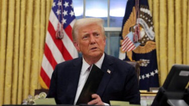 Donald Trump sitting at the Resolute Desk in the Oval Office holding a black folder with flags in the background.