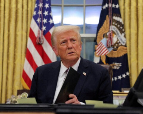 Donald Trump sitting at the Resolute Desk in the Oval Office holding a black folder with flags in the background.