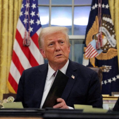 Donald Trump sitting at the Resolute Desk in the Oval Office holding a black folder with flags in the background.