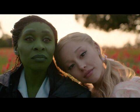 Glinda and Elphaba sit together in a field of red flowers at sunset in a scene from the film “Wicked: For Good,” sharing a quiet moment before unleashing powerful vocals.