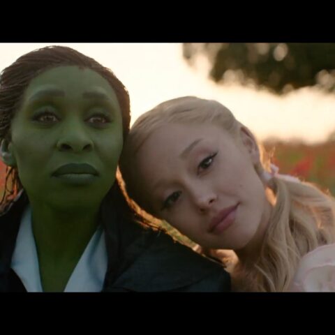 Glinda and Elphaba sit together in a field of red flowers at sunset in a scene from the film “Wicked: For Good,” sharing a quiet moment before unleashing powerful vocals.
