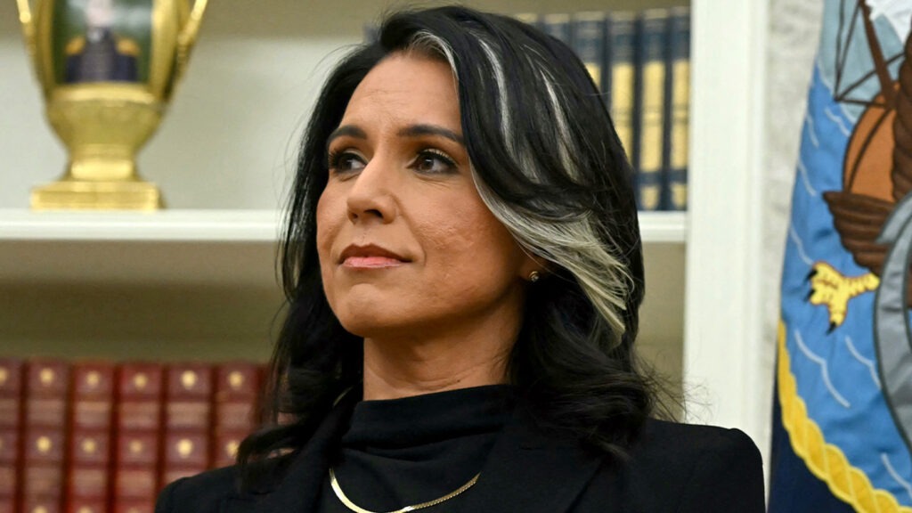 Tulsi Gabbard standing in front of U.S. Navy flag and bookshelf
