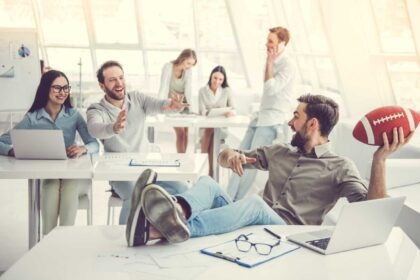 Office coworkers laughing while man holds football at desk.
