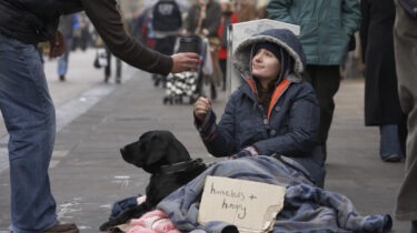 Homeless woman sits on sidewalk with dog while accepting a coffee cup from passerby
