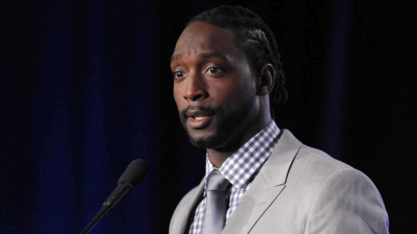 Charles Tillman speaking at a press conference in a gray suit and blue checkered shirt
