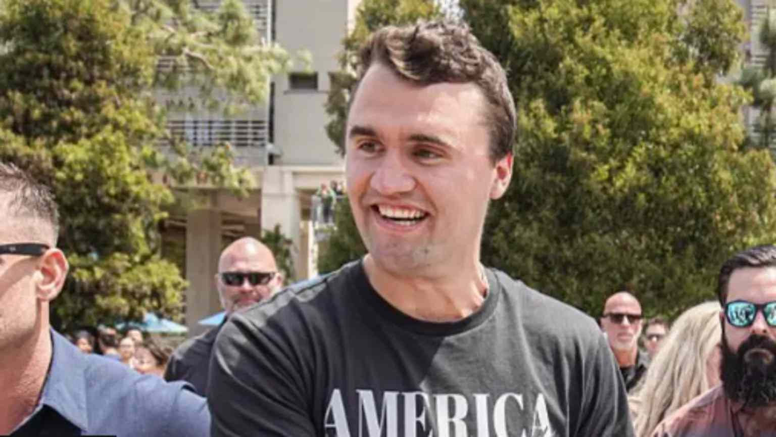 Charlie Kirk smiling at outdoor event surrounded by supporters
