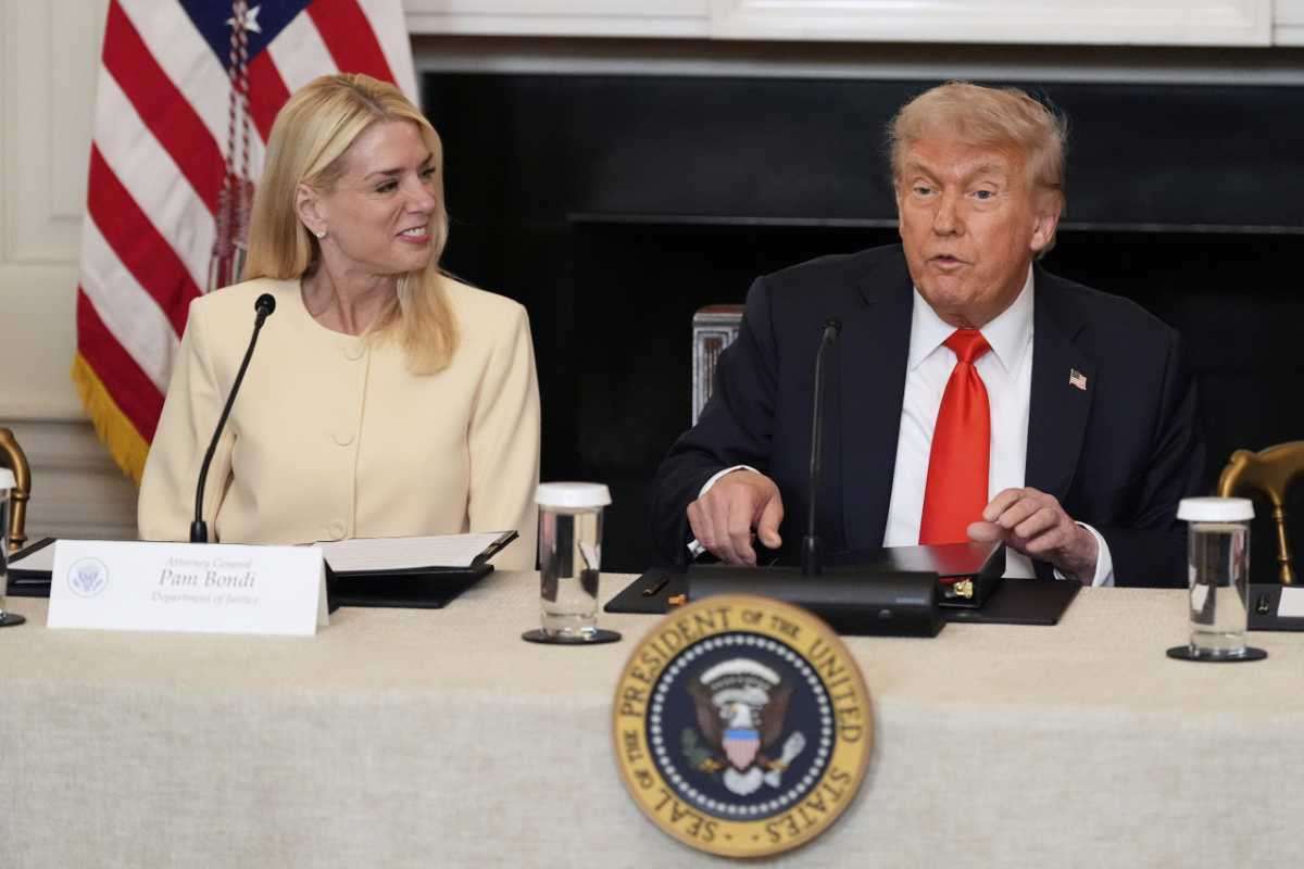 Pam Bondi and President Donald Trump seated at a meeting table with microphones and documents.