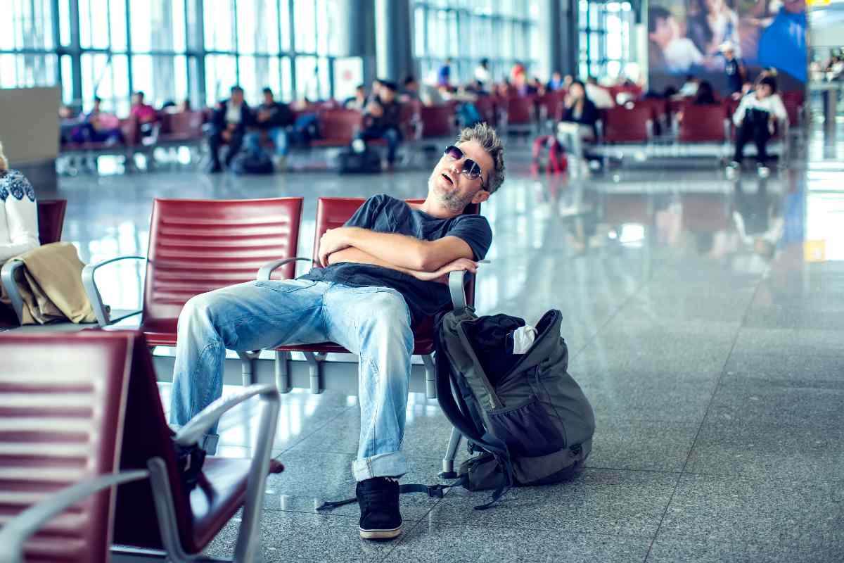 A man sleeping in an airport terminal chair with a backpack beside him.